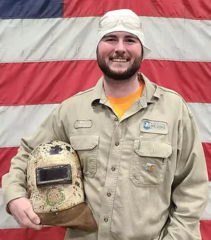 Smiling man in tan uniform holding a worn welding helmet.