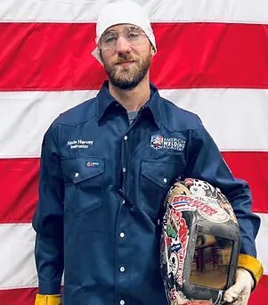 Man in welding gear holding a decorated helmet in front of an American flag.