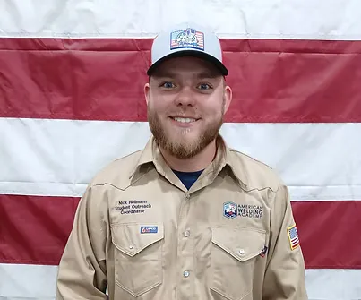 Man in tan uniform and cap with “American Welding Academy” logo, smiling in front of flag.