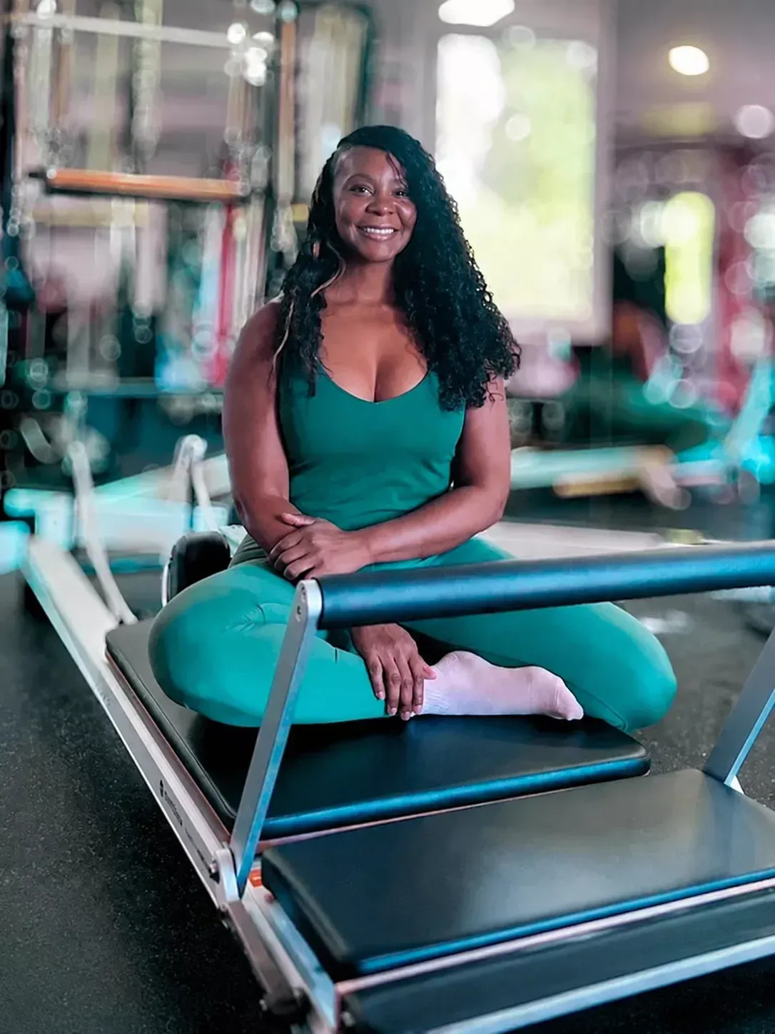 Smiling woman with curly hair sitting on a blue Pilates reformer