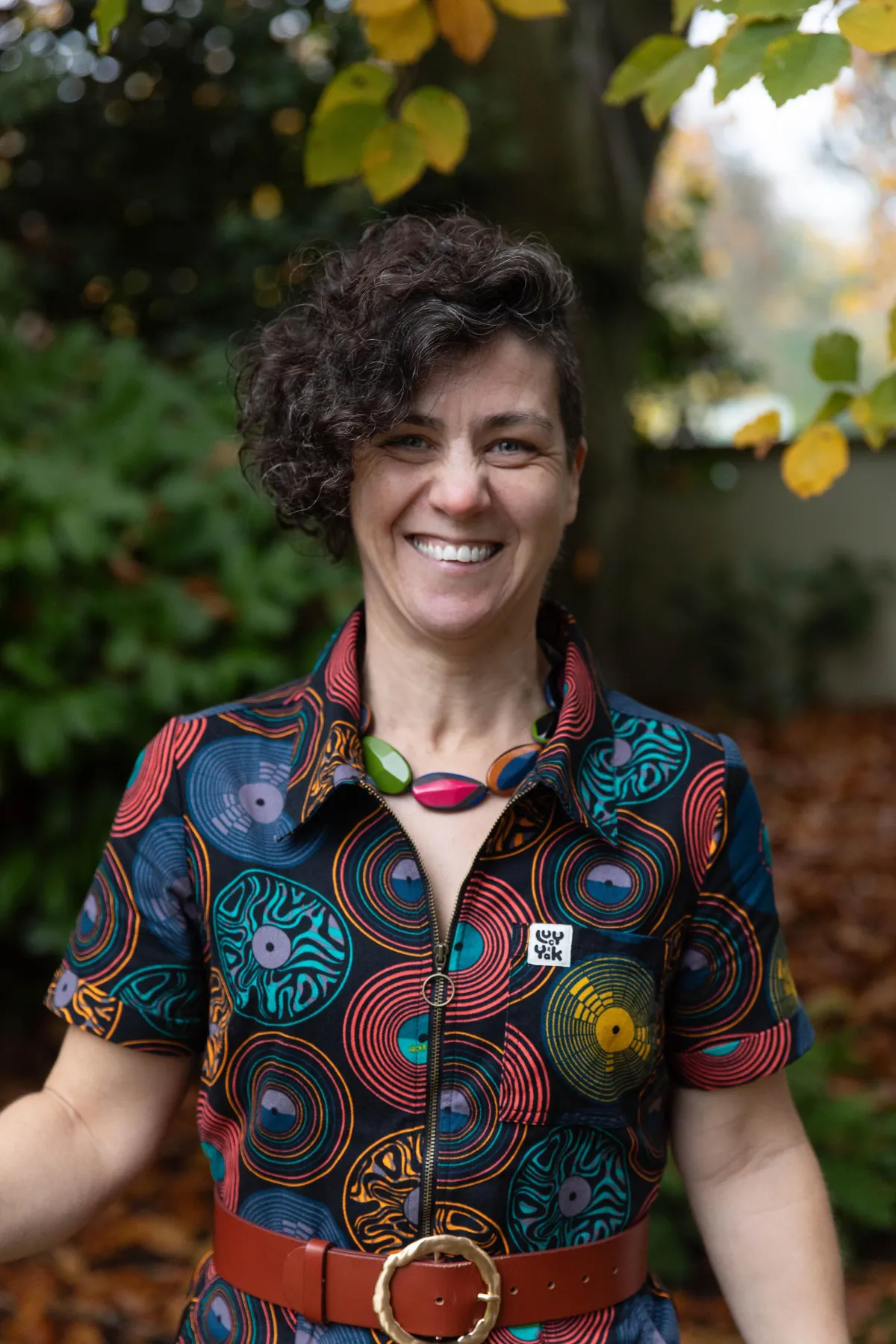 Our founder (a white woman) stands facing the camera, with her arms open and smiling. She supports autistic young people in the north east within their schools and families
