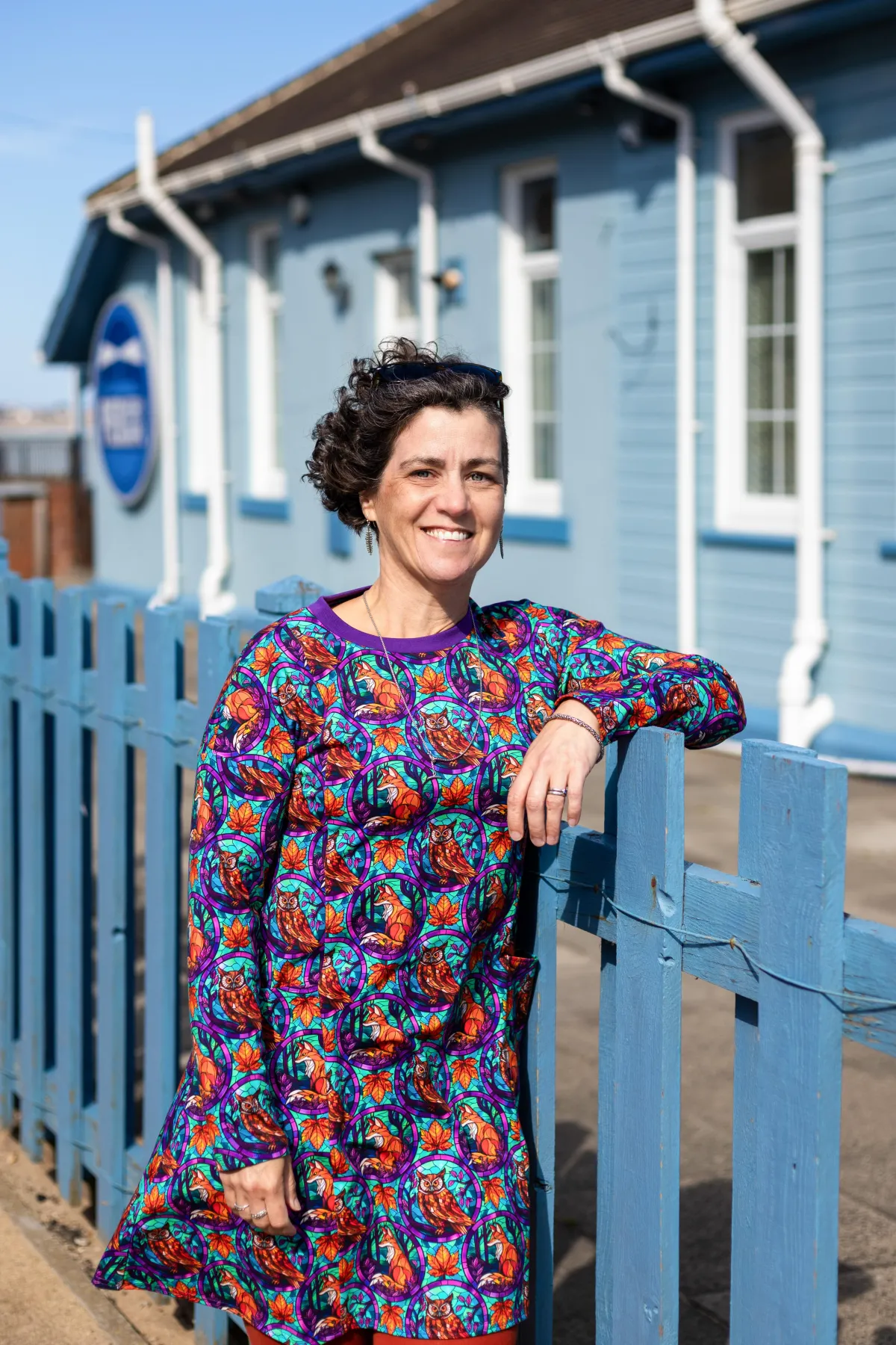 Charlotte (a white woman) is standing in the sunshine smiling at the camera whilst leaning on a fence. She supports autistic young people in the north east within their schools and families