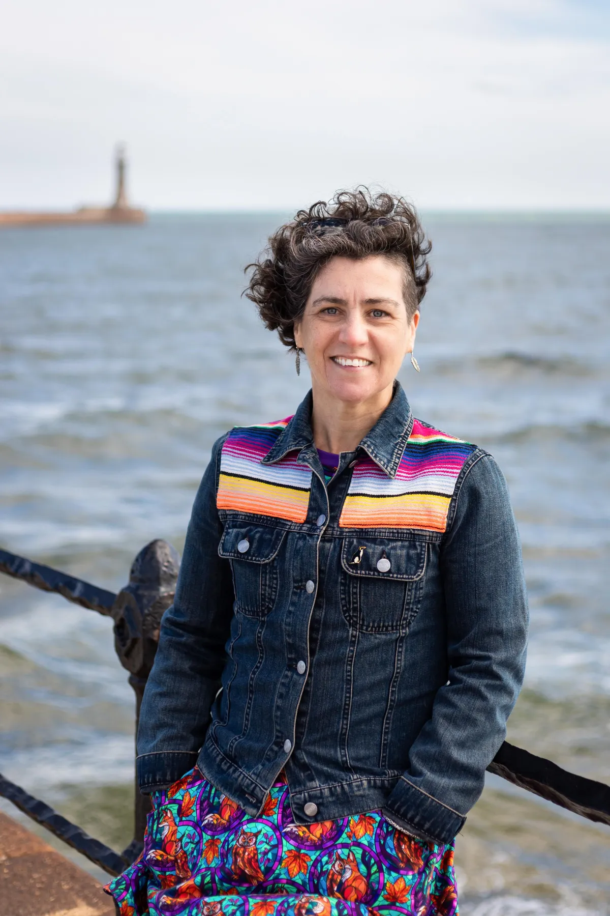 Our founder (a white woman) stands facing the camera and smiling. She is leaning on a railing with the sea behind. She supports autistic young people in the north east within their schools and families