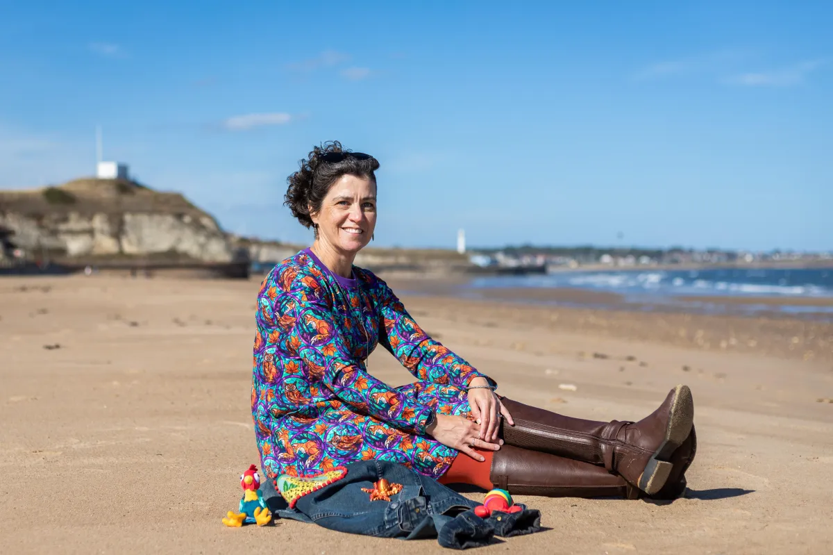 Charlotte (a white woman) sits sideways on to the camera on a beach with the sea behind. She supports autistic young people in the north east within their schools and families