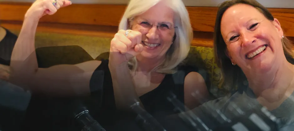 Two women showing their muscles with a dumbbell rack faded in front