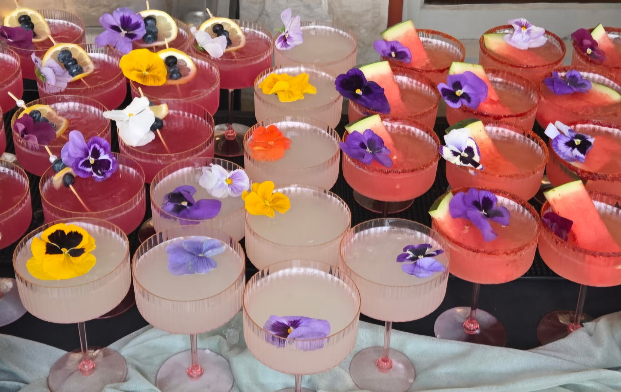 Rows of signature cocktails in coupe glasses garnished with edible flowers for a wedding bar service
