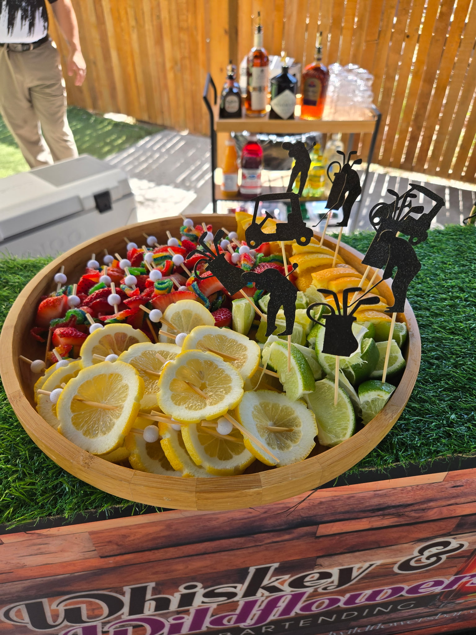 Golf-themed garnish tray with citrus and fruit skewers beside a premium liquor cart at an outdoor golf event