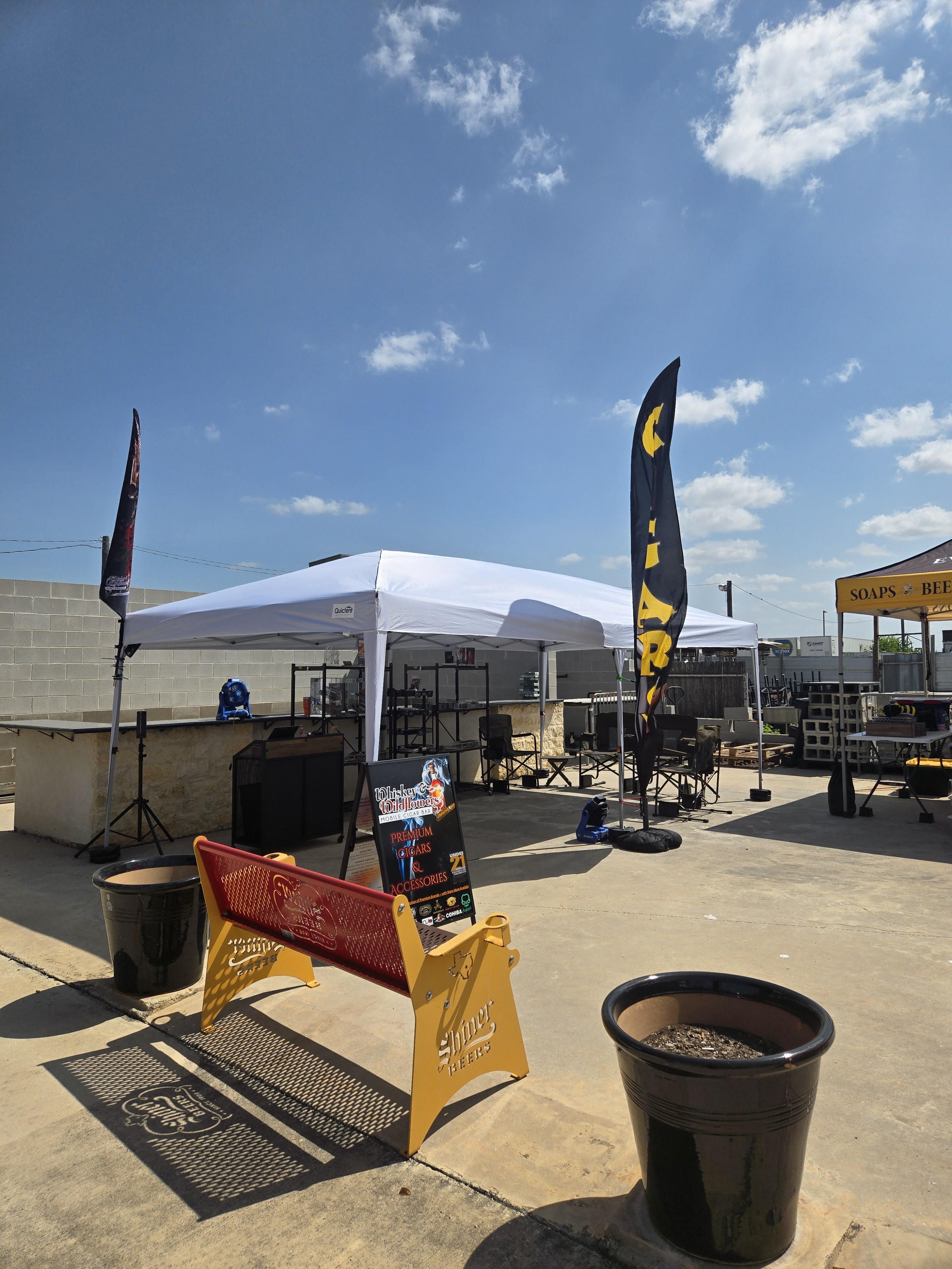 Whiskey and Wildflowers cigar bar tent at an outdoor pop-up market with vendor flags