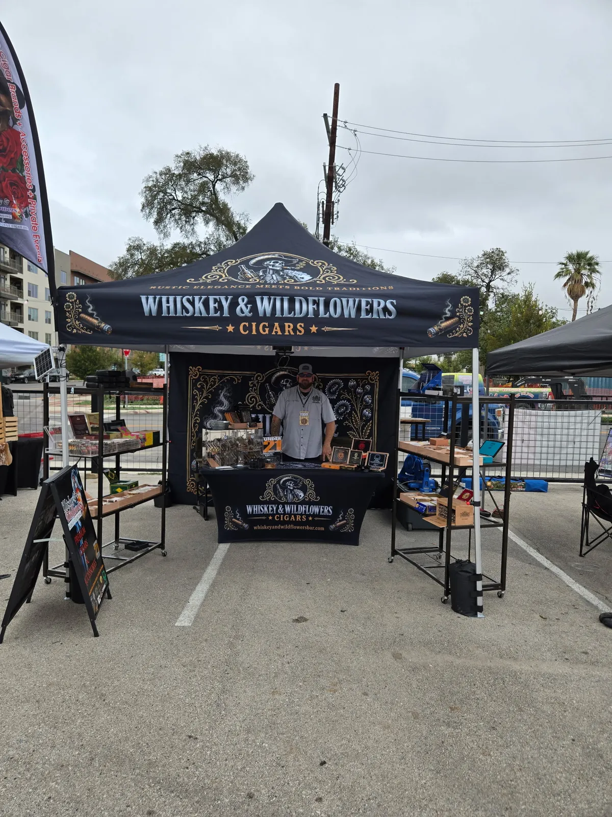 Whiskey and Wildflowers cigar bar tent at an outdoor pop-up market with vendor flags