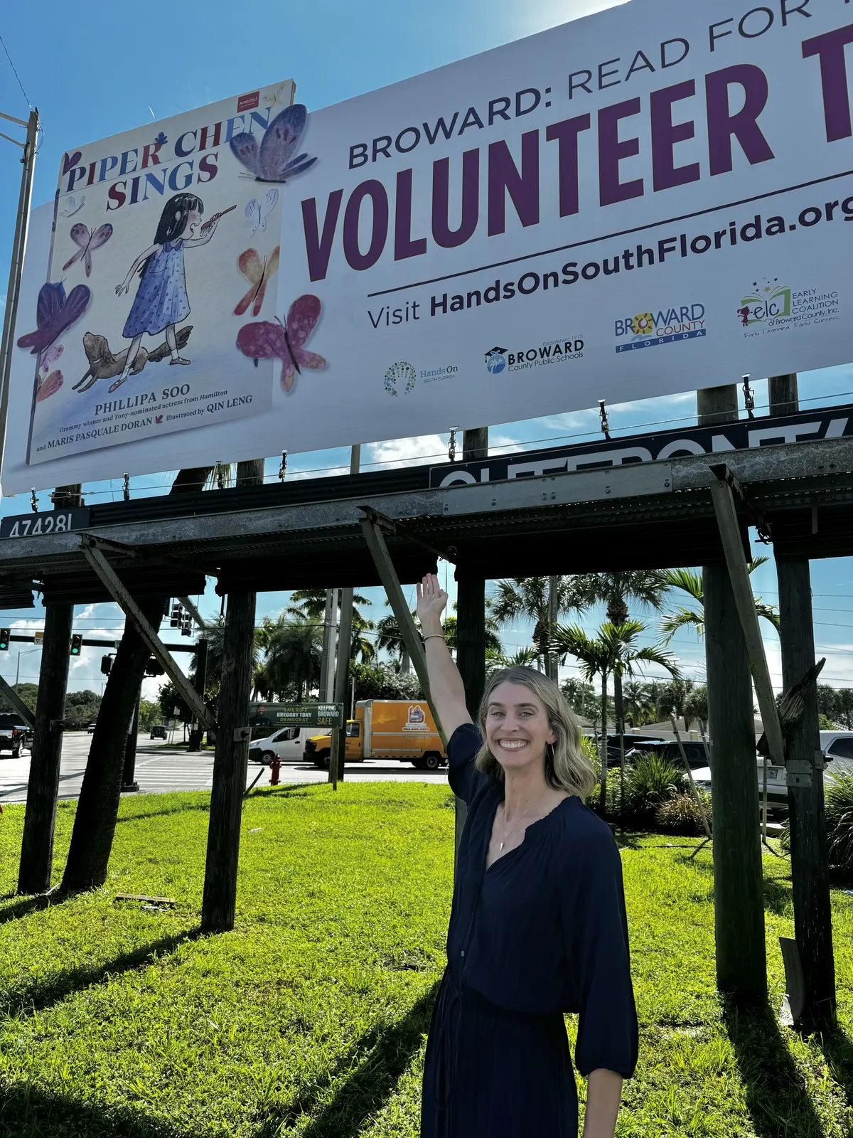 Maris Pasquale Doran pointing up at a Read for the Record billboard featuring Piper Chen artwork.