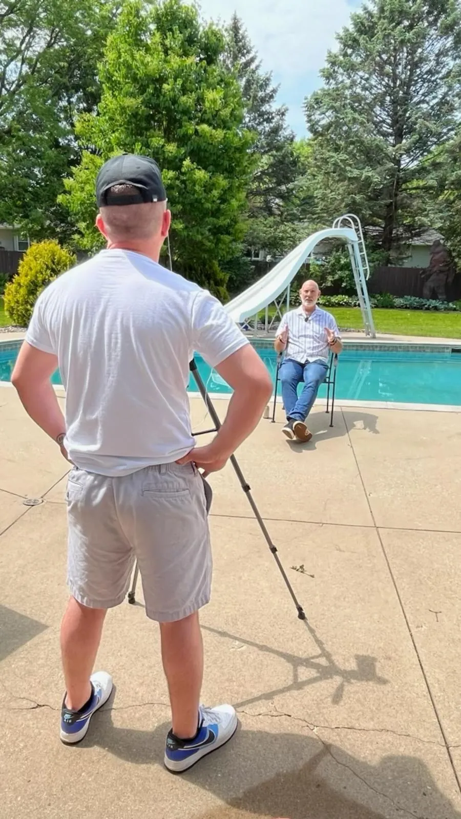 Person filming an interview outdoors by a pool, capturing content for podcasting or social media, with a seated interviewee discussing expertise.