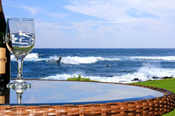 A wide lanai with glass railings overlooking rolling ocean waves, palm trees swaying, and a golden sunset sky. The setting is tranquil, with a small table and two chairs for morning coffee.