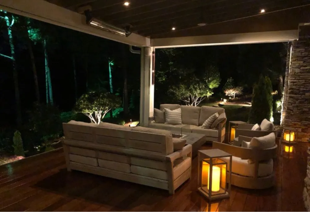 A well-kept backyard with a spacious covered patio featuring retractable Defender Motorized screens attached to a modern house, showcasing white pillars and a lush green lawn under a clear blue sky.