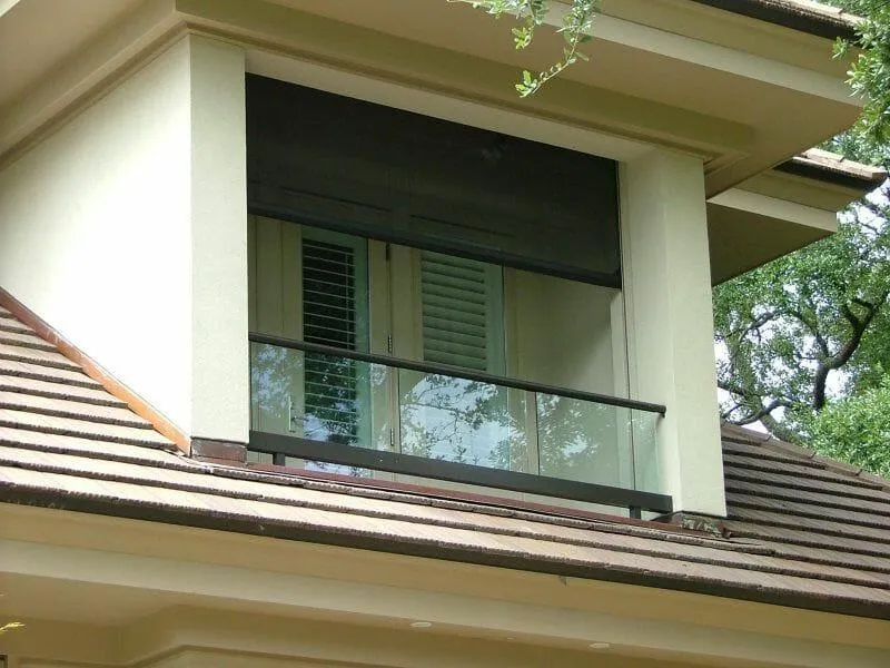 A two-story house with a brick facade, tiled roof, cooper gutters, and landscaped garden featuring a palm tree and retractable Motorized Defender Screens on the windows. There's a black SUV parked on the driveway.