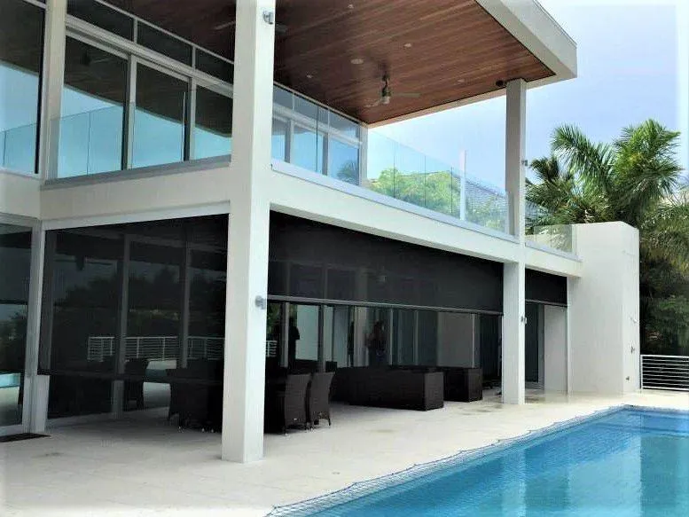 Modern two-story home with large windows, a wooden ceiling overhang, an outdoor seating area equipped with a retractable insect screen, and an adjacent swimming pool surrounded by palm trees.