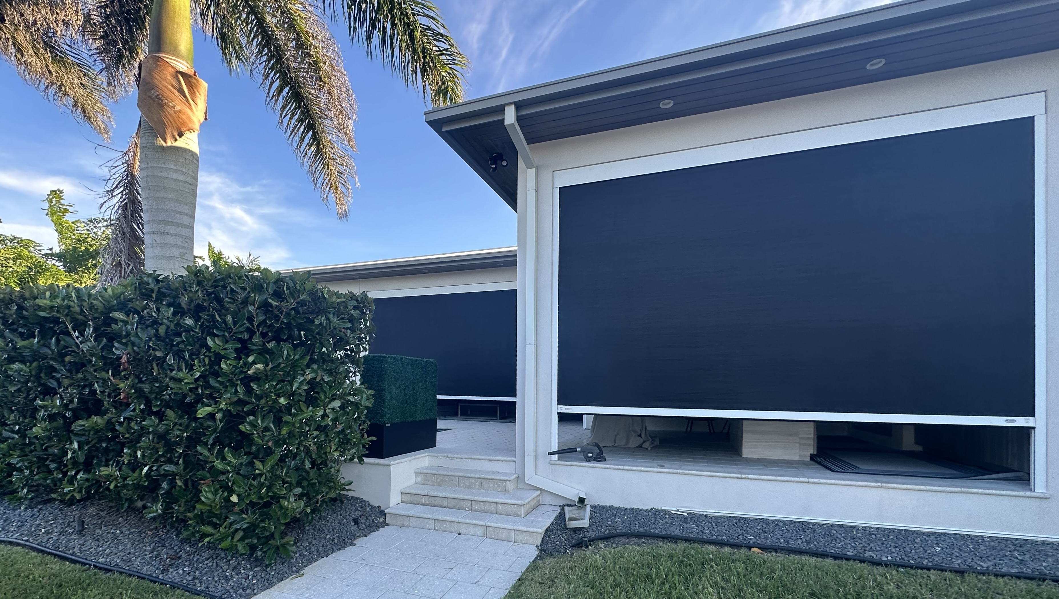 A photo of a white house with two retractable privacy Screens.  The second screen sit deaper in the house stucture then the first. A tall Royal Queen Palm in front. 