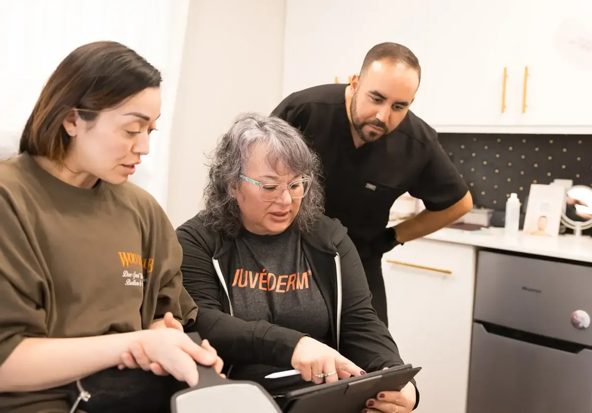 Three aesthetic professionals review information on a tablet together, with one woman pointing at the screen in a clinical office.