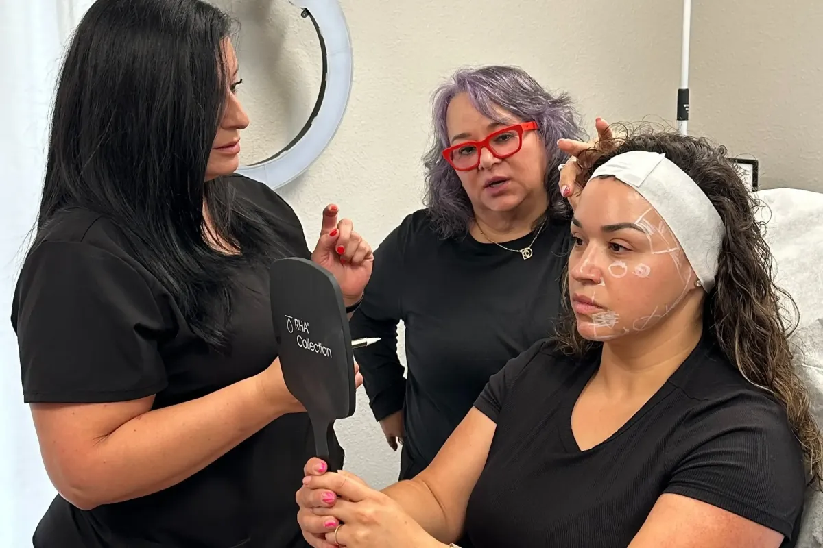Practitioner holds a mirror for a patient with white facial injection markings while a colleague with red glasses explains the upcoming treatment.