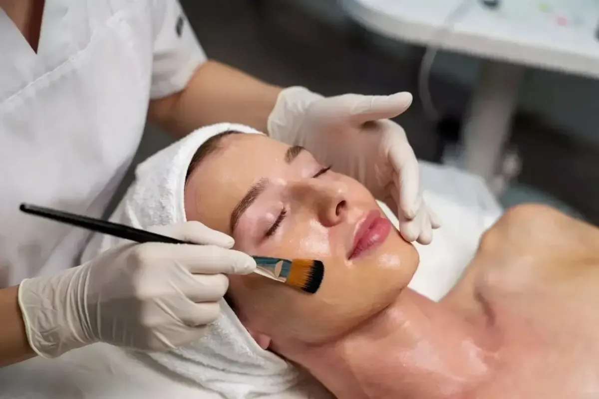 Gloved aesthetician applies a chemical peel solution to a patient’s face with a brush during a professional treatment in a clinical setting.