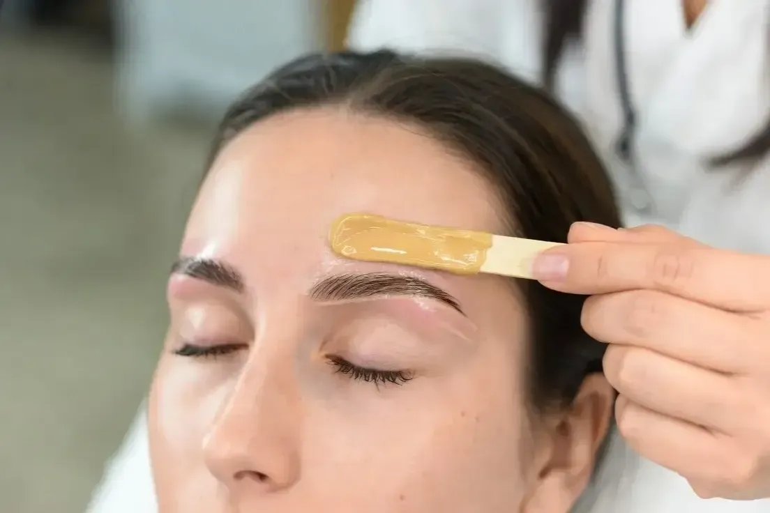 Practitioner applies wax to a woman’s eyebrow using a wooden spatula during a professional brow wax treatment.