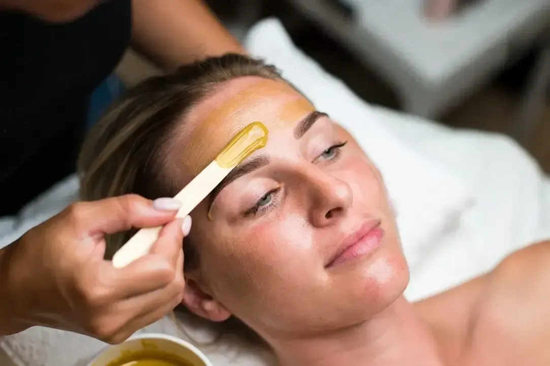 Practitioner applies wax to a woman’s forehead during a full face wax treatment while she lies relaxed on a treatment bed.