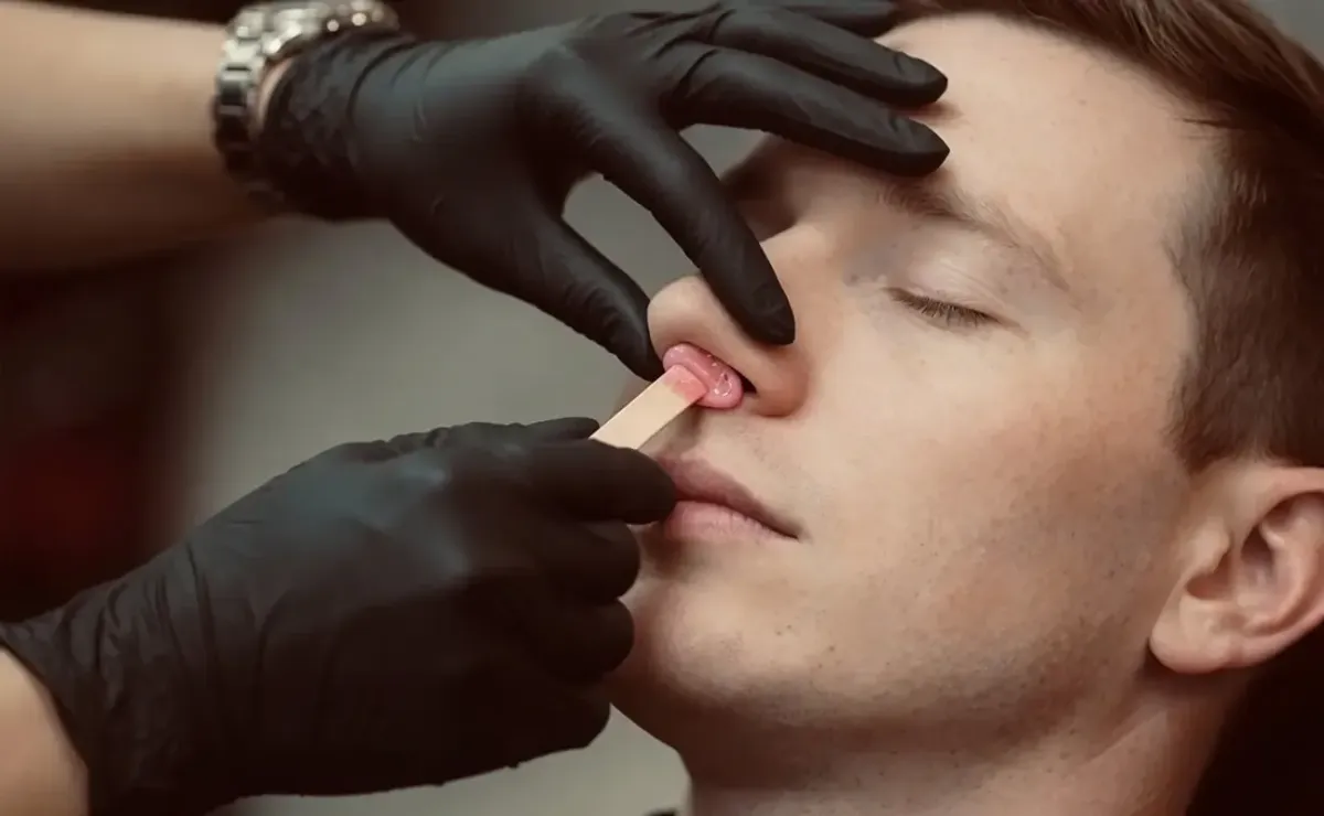 Gloved practitioner applies wax inside a man’s nostril during a nose wax treatment while he lies with eyes closed.