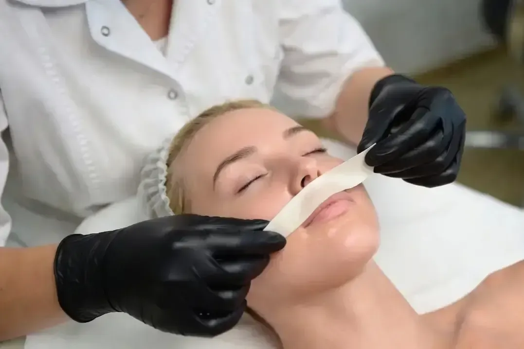 Gloved practitioner applies a wax strip to a woman’s upper lip during a lip wax treatment while she lies with eyes closed.