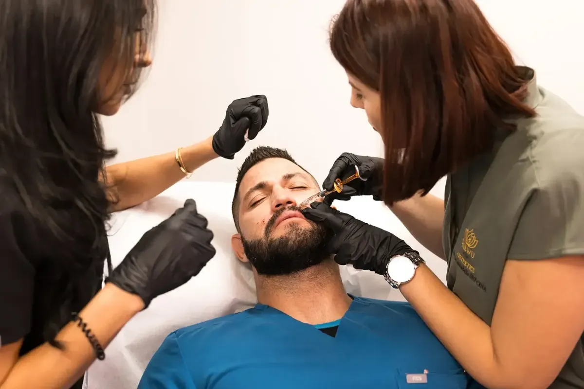 Two gloved medical professionals administer a cosmetic injection to the face of a relaxed bearded man lying down with eyes closed.