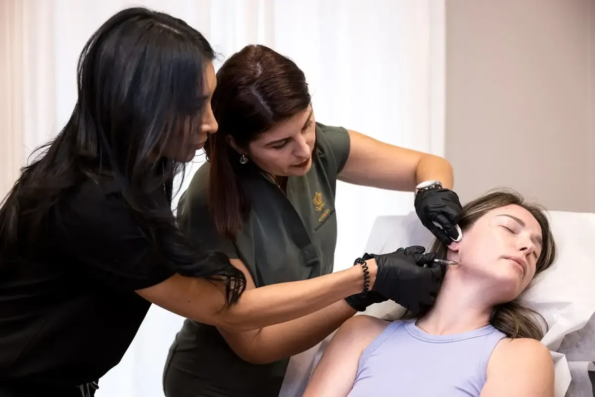 Two gloved medical professionals administer a cosmetic injection to the chin of a relaxed young woman lying down with eyes closed.