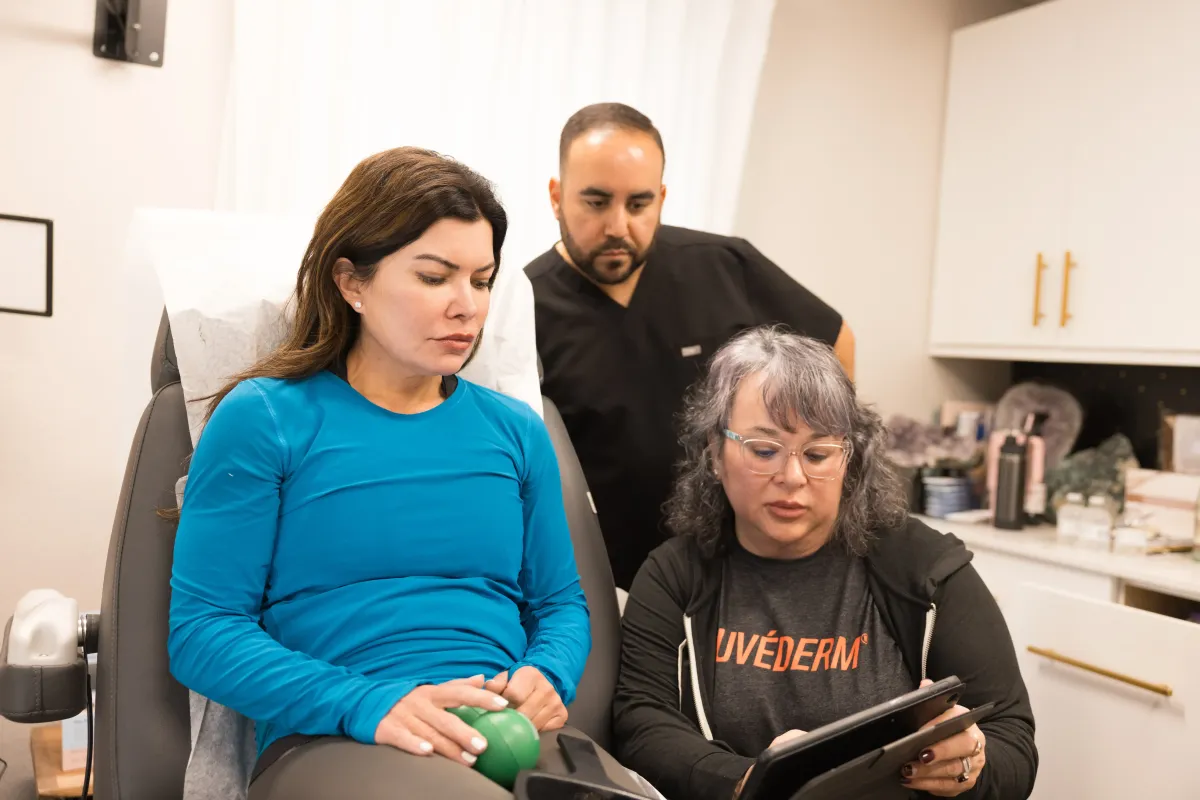 Patient listen to a laser hair reduction consultation while two providers review her treatment plan on a tablet in a clinical setting.