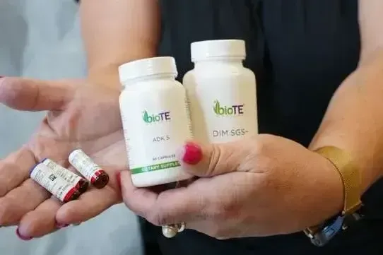 Person holding BioTE ADK 5 and DIM SGS+ supplement bottles with capsules in their hands, showcasing hormone support dietary supplements.