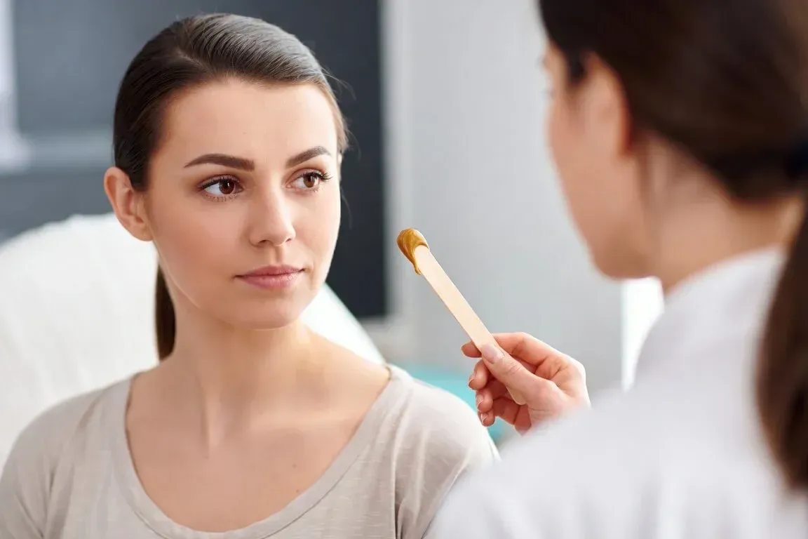 Aesthetician holds a wooden spatula with wax during a waxing consultation with a client in a clinical setting at Chrome Aesthetics.