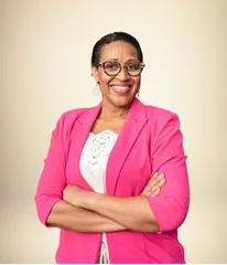  A professional headshot of a smiling woman with glasses and dark brown hair. She is wearing a bright pink blazer and a white shirt, with her arms crossed confidently. The background is a soft, neutral beige.
