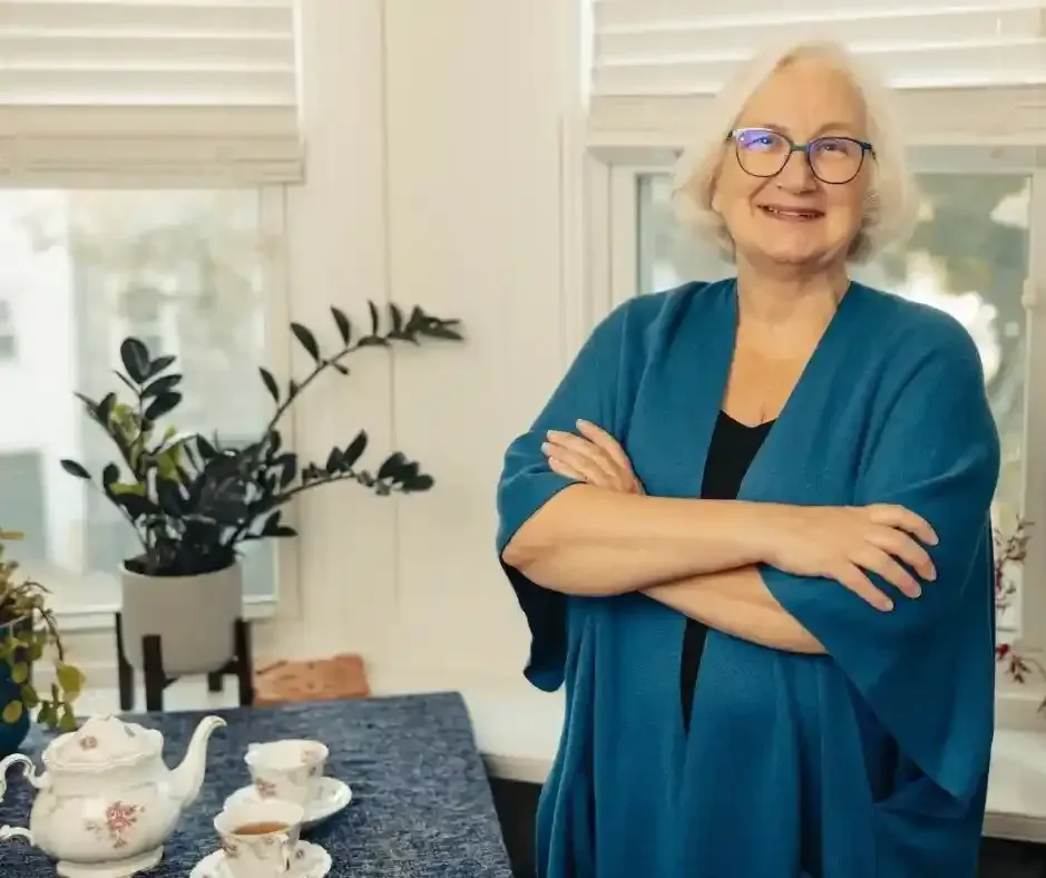 Smiling older woman standing with arms crossed in a bright room with plants and tea set, representing a calm hypnotherapy practitioner