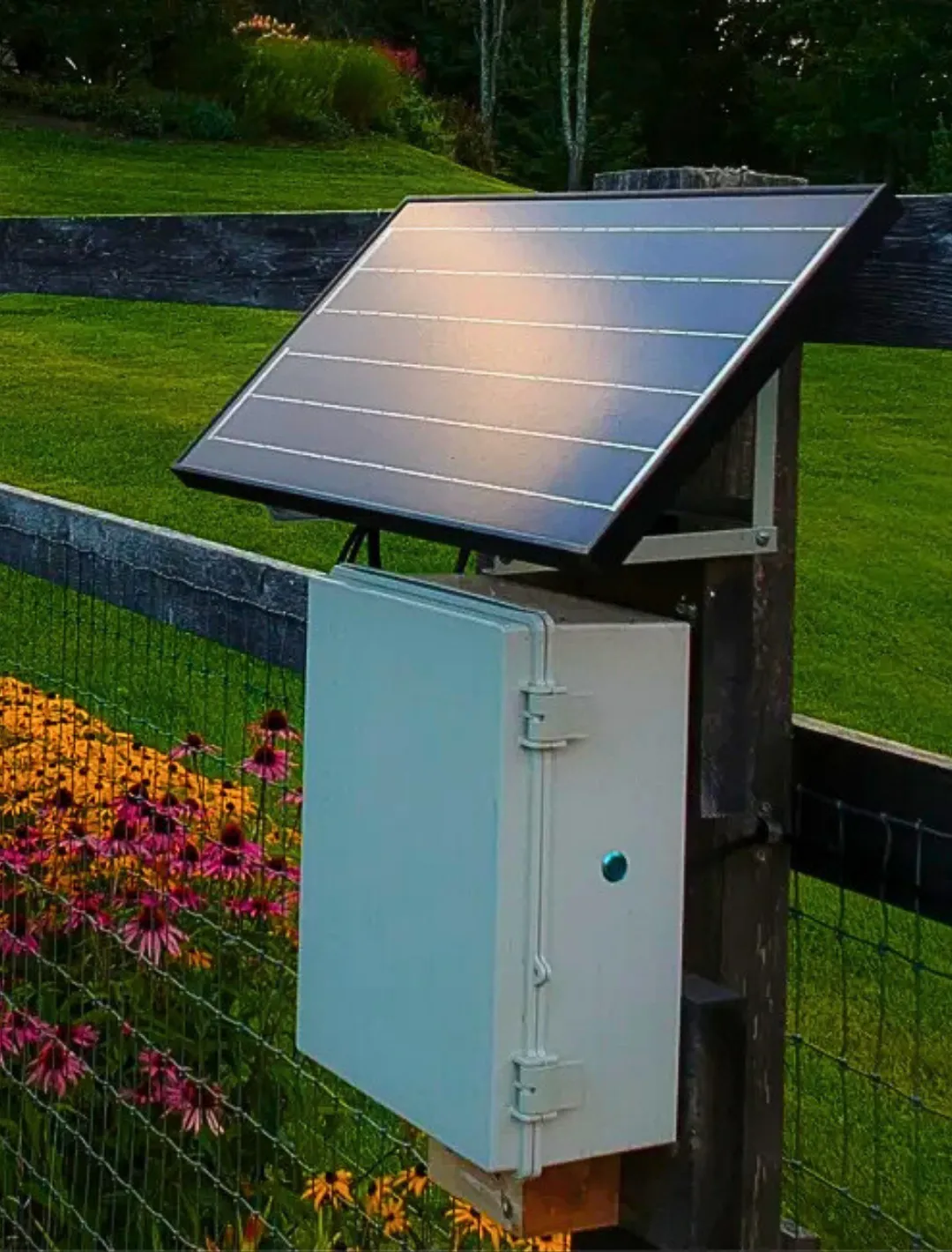 Boîtier principal du système SentiControl avec panneau solaire, conçu pour contrôler l'irrigation et l'éclairage de plusieurs zones, fixé sur une clôture entourée de fleurs dans un jardin.