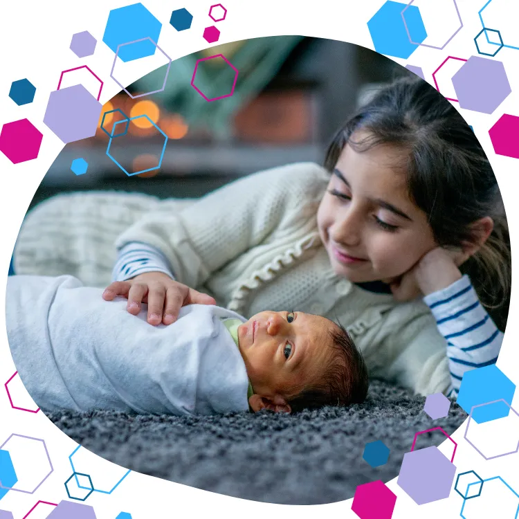 newborn baby lying swaddled on the floor whilst older sister is lying next to them, with a hand on their chest. CubCare refresher antenatal classes.