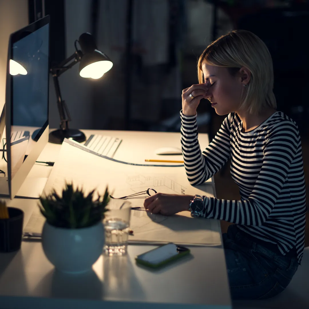 A woman sitting at her desk with her laptop screen on. She is a wedding professional and is tired, frustrated, likely has a headache She has blonde hair and a white and black striped, long sleeve shirt on with blue jeans. Room lighting is dim. There is a desk lamp turned on, creating a feeling of late night work. The desk has paper strewn about, a succulent plant in a white bowl, iphone, glass of water, eye glasses, a pen and pencil on the deks.