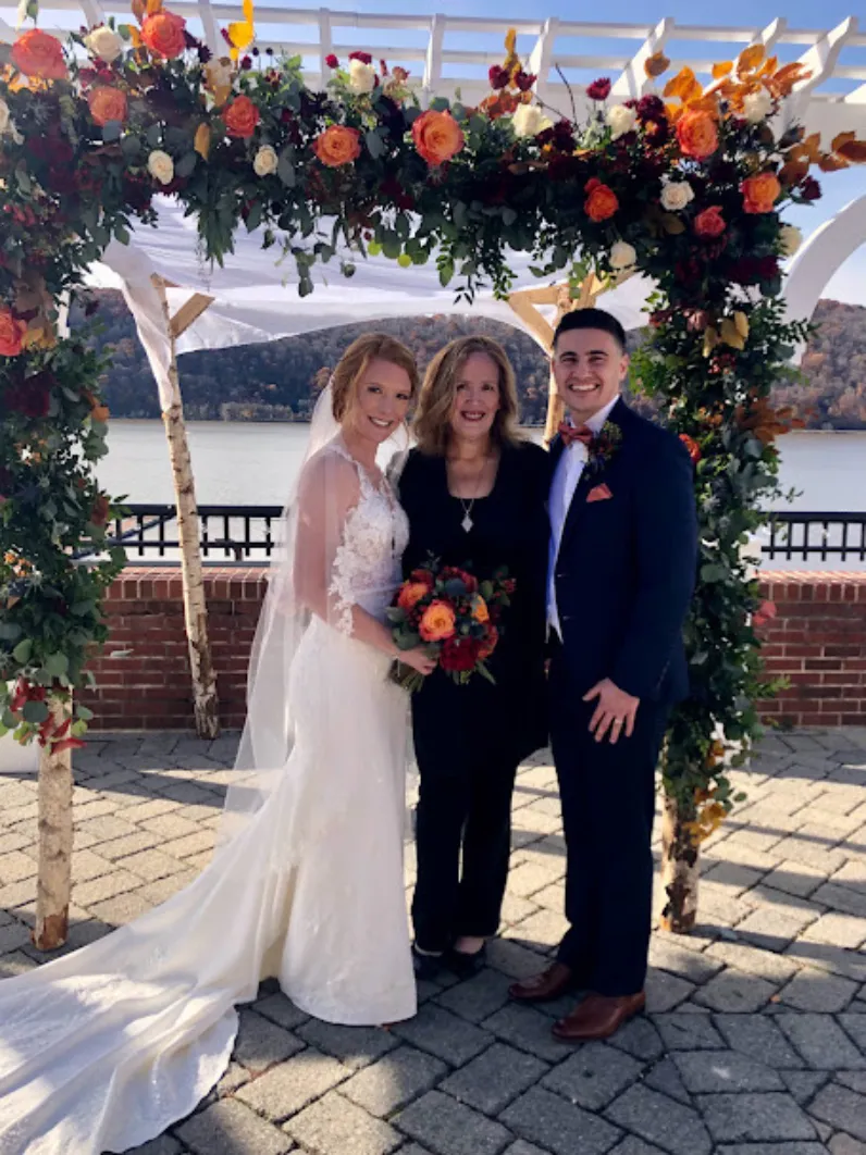 Lisa Zaro, Professional Officiant with couple after ceremony at the Grandview in Poughkeepsie, NY. Pictured (L) Bride, (C) Lisa Zaro, (R) Groom. Archway is adorned with peach, white, yellow and pink roses, plus greenery. The Hudson River and western coast of the river can be seen in the background.