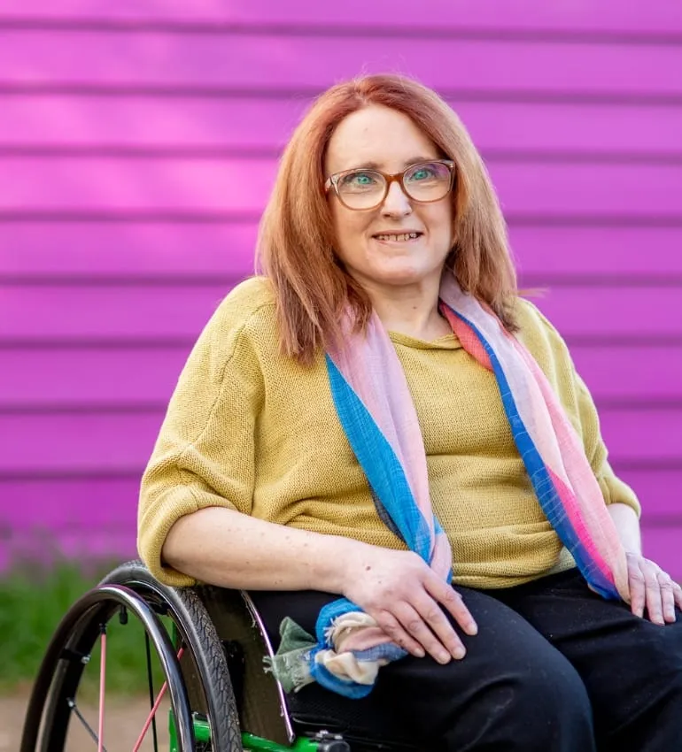 Ainslee Hooper smiling, seated in her wheelchair in front of a pink wall, wearing a mustard top and colourful scarf.