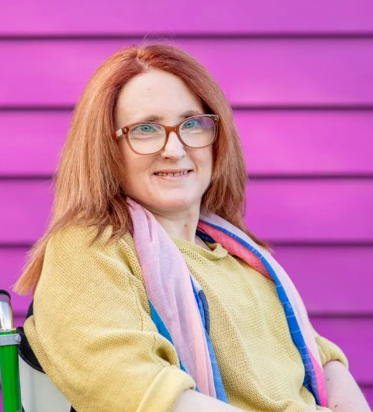 Ainslee Hooper smiling in front of a pink wall, wearing a mustard top and colourful scarf.