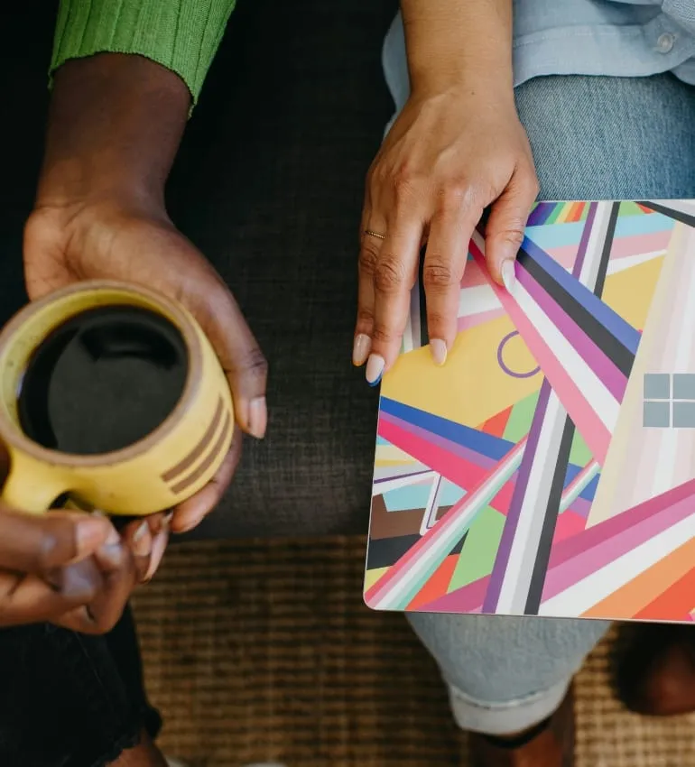 Two people seated together, one holding a coffee cup and the other holding a colourful notebook.