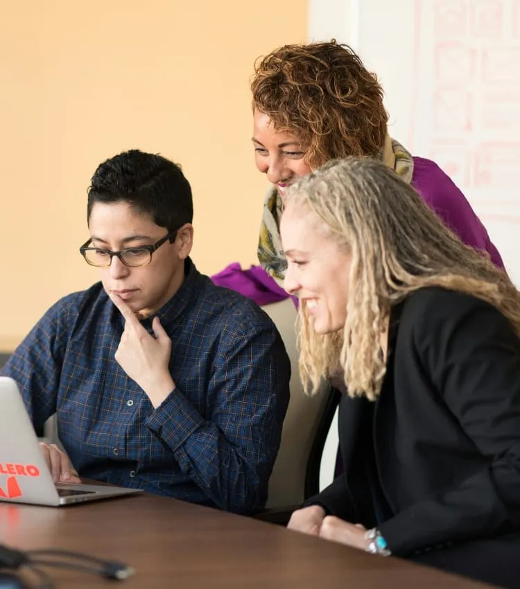 Three colleagues gathered around a laptop, smiling as they work together.