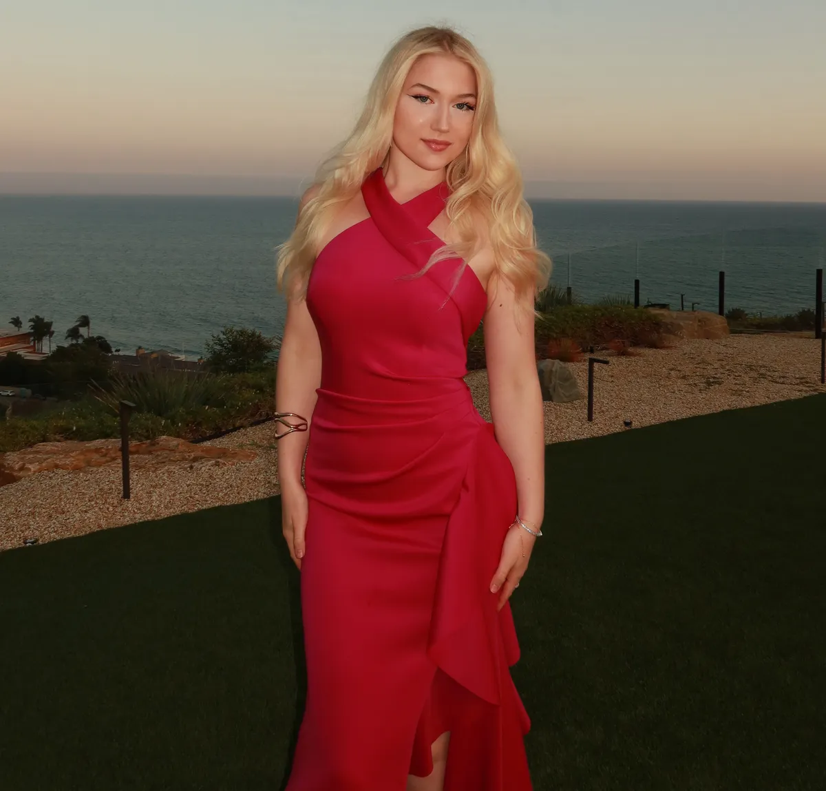 Portrait of a blonde woman in a gold ruched dress looking off-camera against a dark red backdrop, framed in an arched window shape.