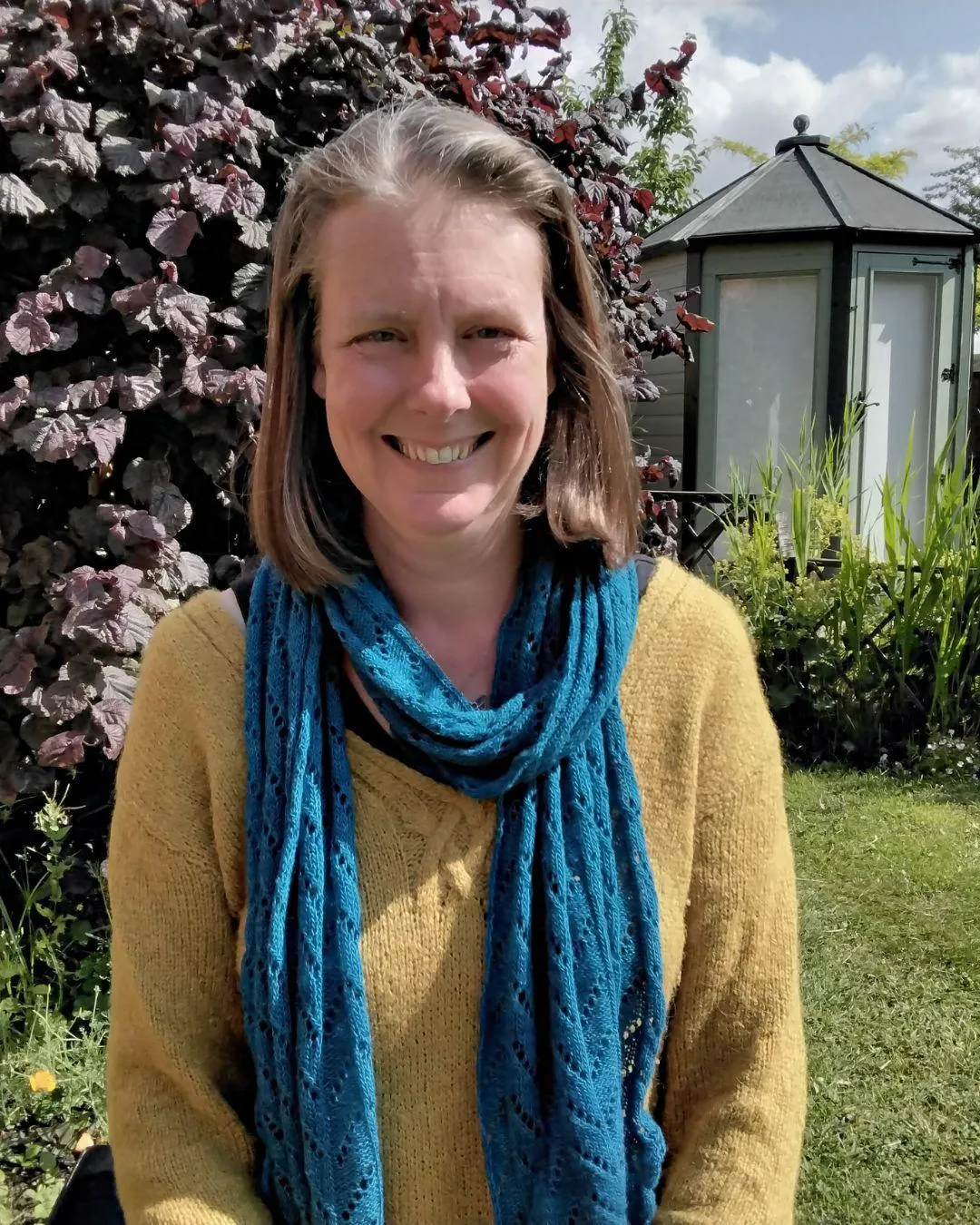 Woman wearing a yellow sweater and a blue scarf standing in a garden (Jodie Clark).