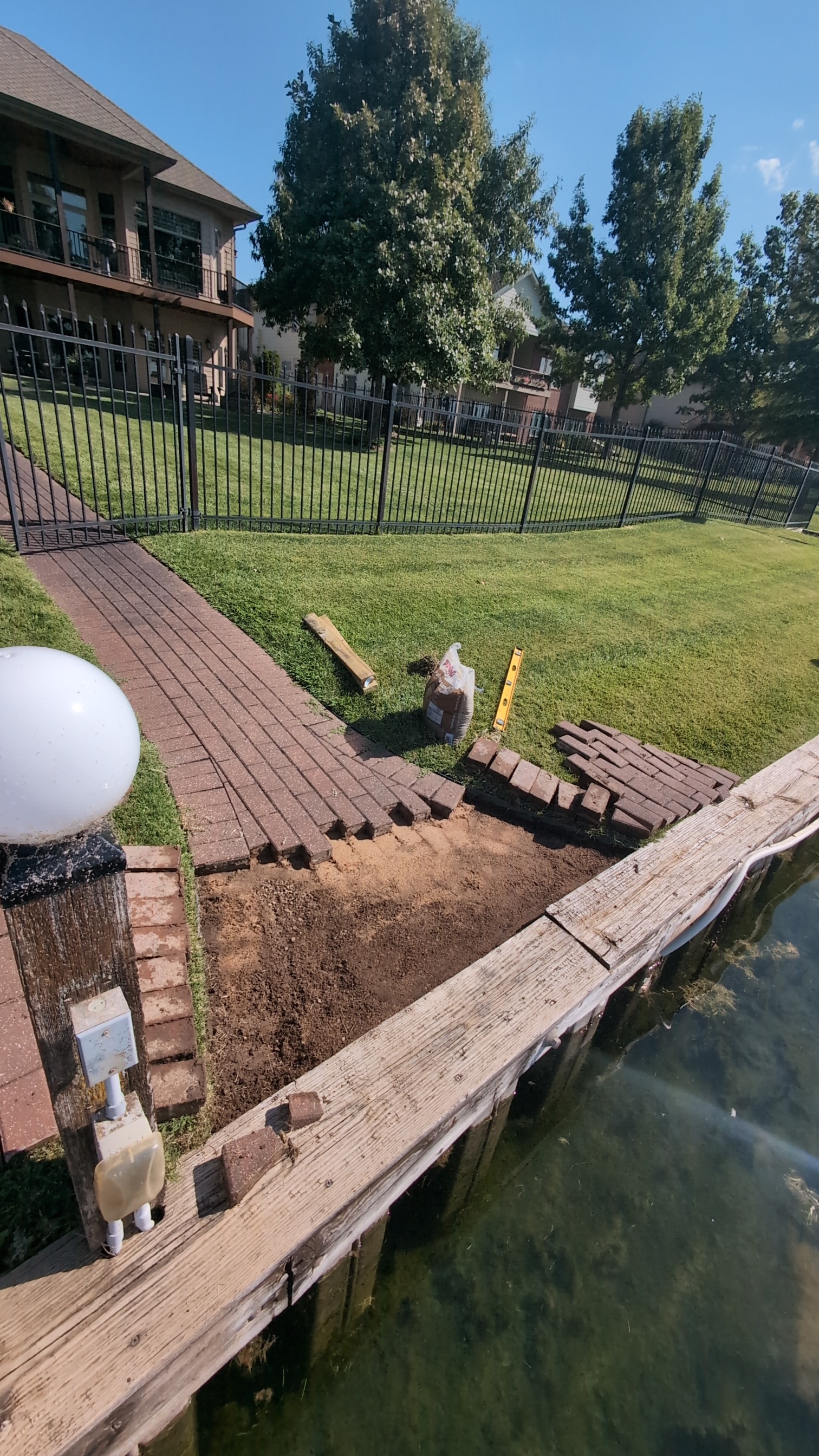 Paver walkway installation in progress near a waterfront, featuring a partially completed pathway, landscaping elements, and tools for handyman services.