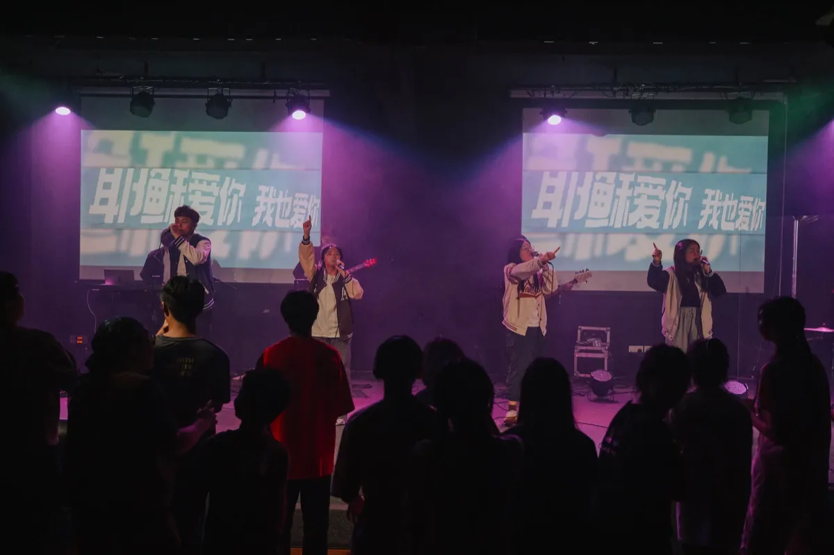 过往教会青年活动中，多位表演者在紫色灯光的舞台上演唱，前景可见观众。