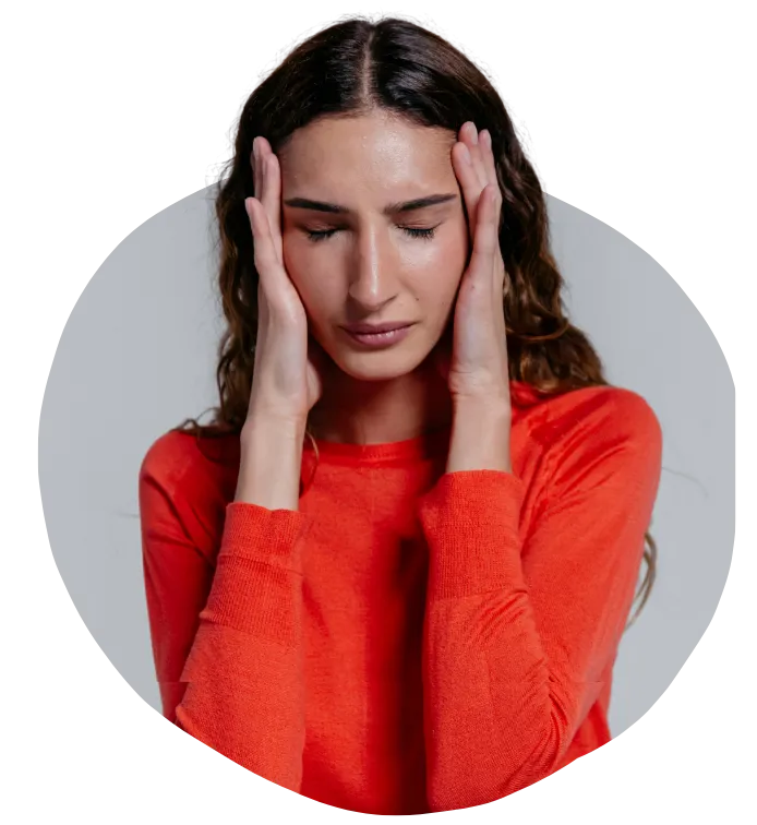 A woman  long, wavy brown hair wearing a bright orange sweater is shown with her eyes closed and her hands pressed against her temples. She appears to be in pain or experiencing a headache.
