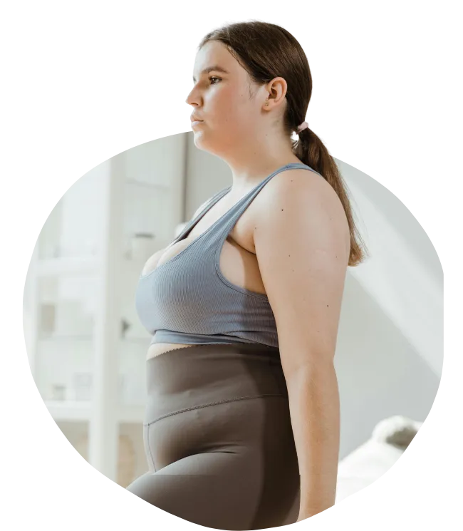 A profile view of a woman with her hair in a ponytail, wearing a blue sports bra and grey high-waisted leggings. She is standing indoors in a bright, minimalist room with a white shelf in the background.
