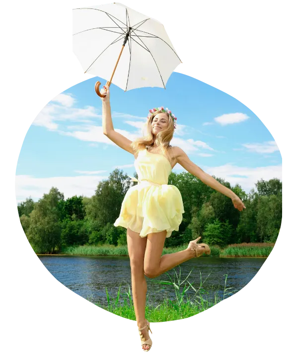 A woman in a light yellow dress leaping playfully in a grassy area near a lake. She is holding a white umbrella high in her right hand and wearing a floral headband.