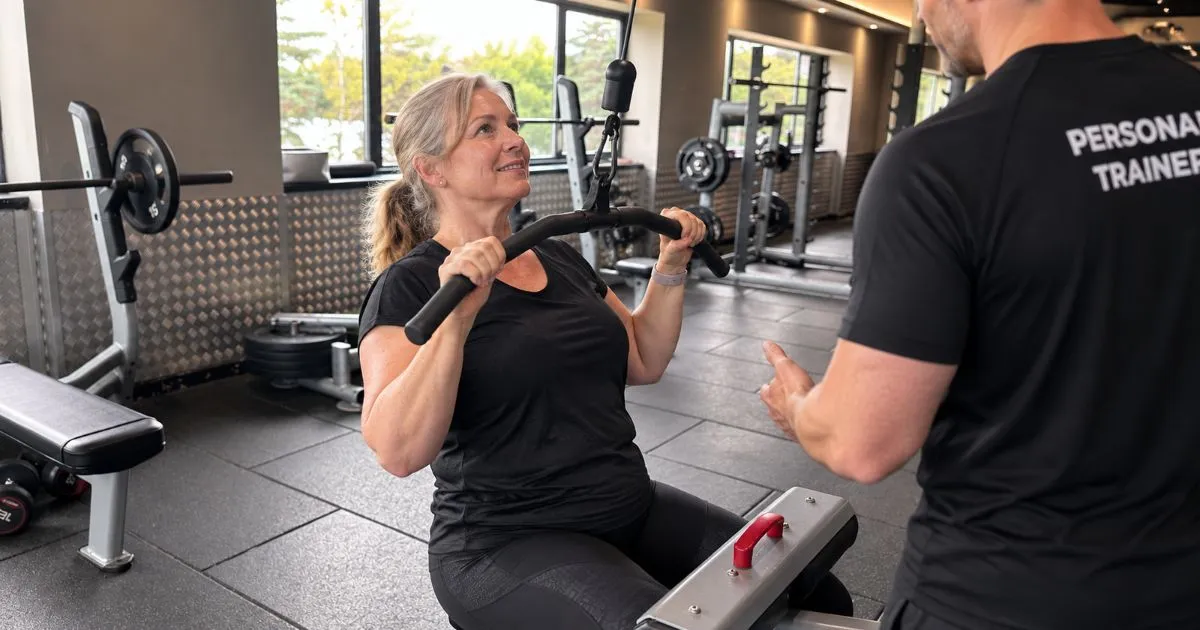 Personal trainer Rob Ede coaching a smiling woman in her 50s on the lat pulldown machine at Lightwater Leisure Centre